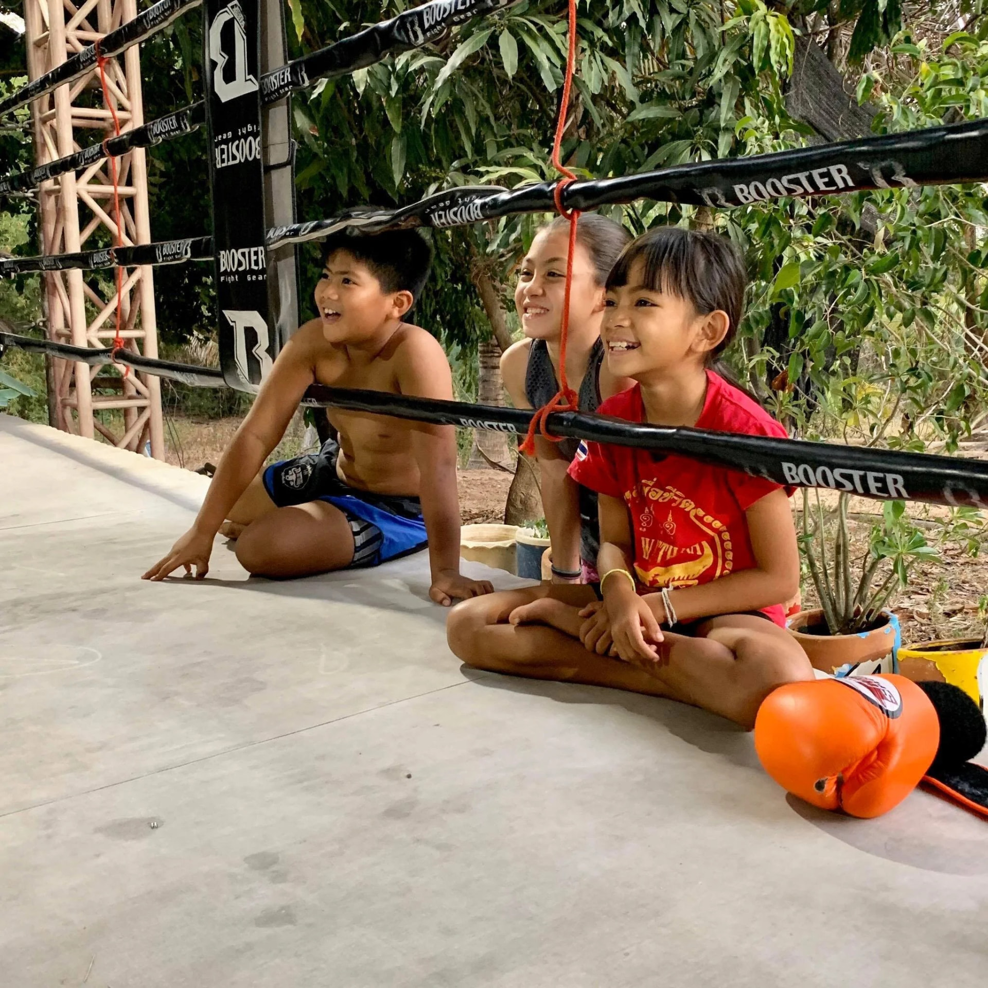 Three children sitting under the ring ropes, laughing as they watch training.