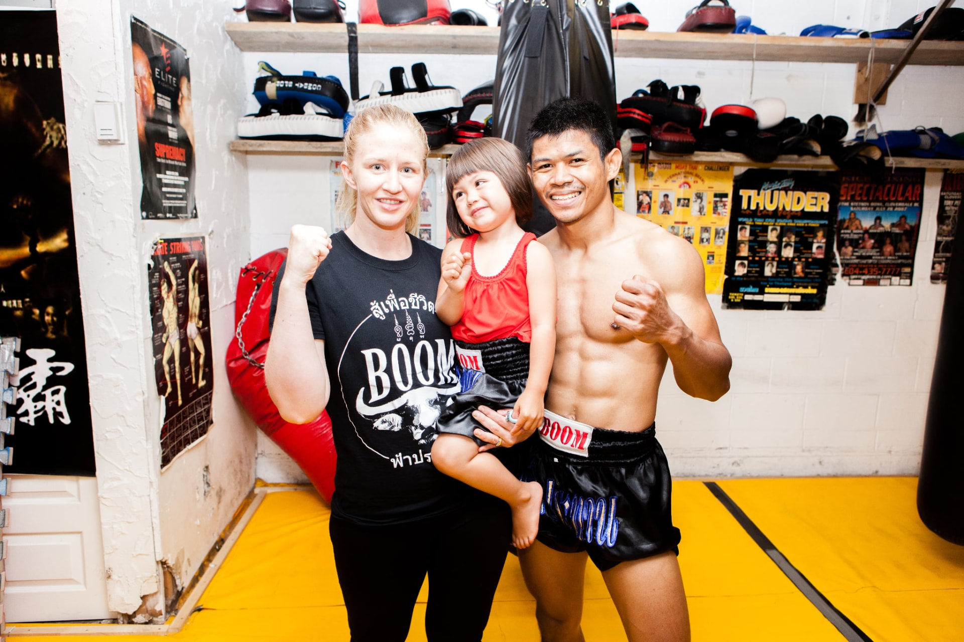 Frances and Boom standing together with their young daughter inside a Muay Thai gym.