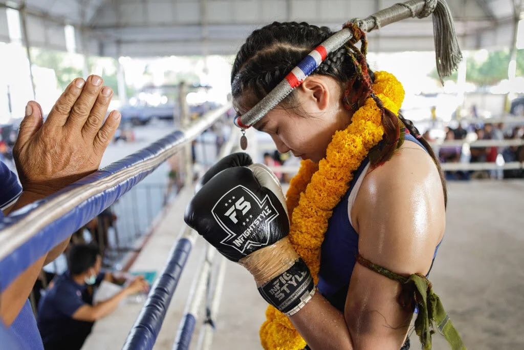 A young fighter in a flower garland lowering her head in prayer while a coach stands at the ropes.